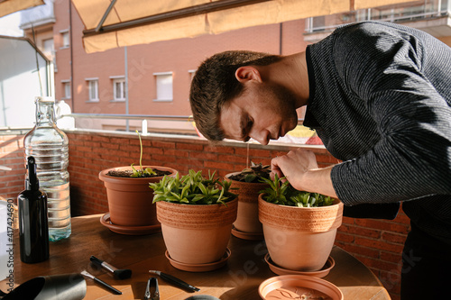 Hombre joven caucásico cuidando varias de sus plantas en la terraza con variedad de herramientas en la mesa de su casa durante el atardecer.