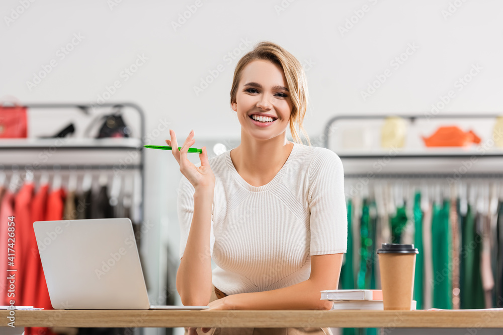 Positive seller holding pen near laptop and coffee to go in showroom Positive seller holding pen near laptop and coffee to go in showroom