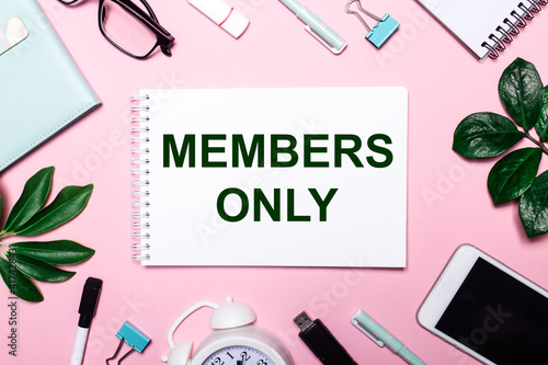 MEMBERS ONLY is written in a white notebook on a pink background surrounded by business accessories and green leaves.