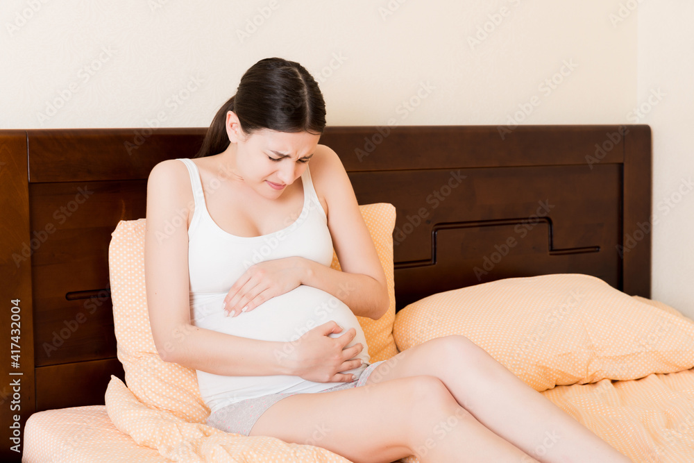 Young pretty pregnant woman, in pain, sitting on bed in bedroom, preparing to give a birth Young pretty pregnant woman, in pain, sitting on bed in bedroom, preparing to give a birth