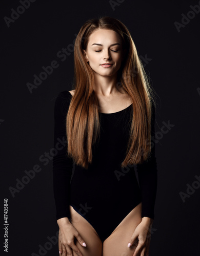 Young beautiful woman with long silky straight hair in black body with eyes closed standing and posing over dark background. Haircare, beauty, wellness concept