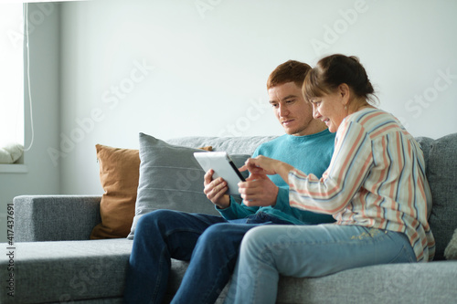 son teaching his mother to use tablet. older people using technology. Cheerful elderly woman sitting on the sofa next to his adult son