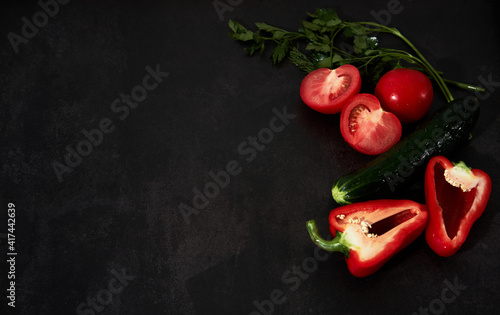 Fresh vegetables on black kitchen table. Minimalistic restaurant menu. Ingredients for salad. Red bell pepper and tomato, green cucumber with greens on black background with space for text.