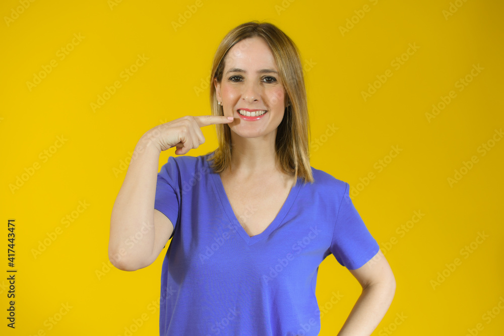 Young beautiful woman wearing t-shirt standing over yellow isolated background smiling cheerful showing and pointing with fingers teeth and mouth. Dental health concept. Young beautiful woman wearing t-shirt standing over yellow isolated background smiling cheerful showing and pointing with fingers teeth and mouth. Dental health concept.