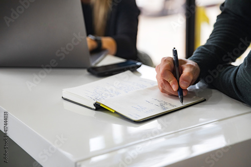 Man writing in notebook next to coworker on laptop and phone in the morning office meeting with a white table and windows