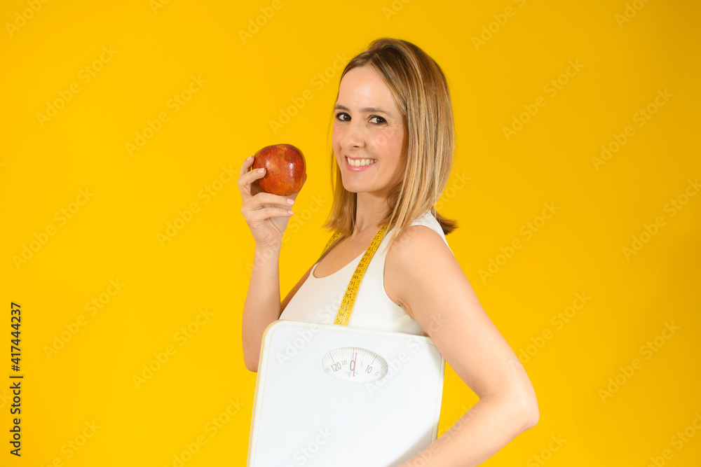 Beautiful woman holding an apple with scales over yellow background Beautiful woman holding an apple with scales over yellow background