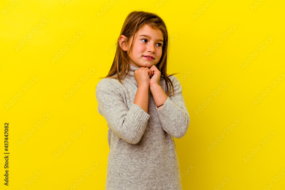 Little caucasian girl isolated on yellow background keeps hands under chin, is looking happily aside. Little caucasian girl isolated on yellow background keeps hands under chin, is looking happily aside.