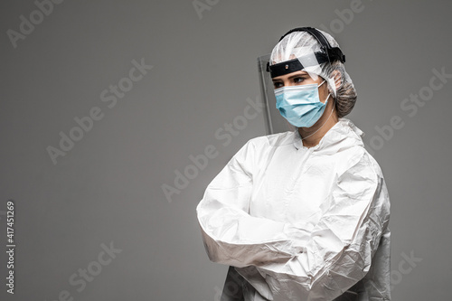 Woman doctor wearing white hazmat suit medical gloves and respirator standing isolated on gray background quarantine crossed hands posing to camera confident