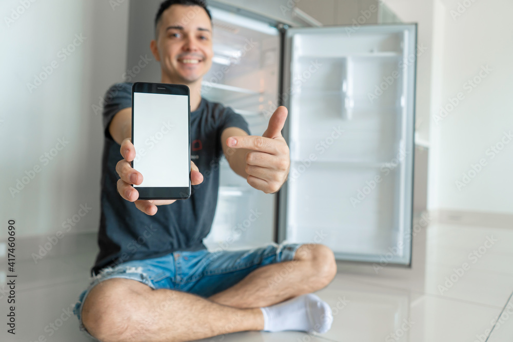 A young guy orders food using a smartphone. Empty refrigerator with no food. Food delivery service advertisement A young guy orders food using a smartphone. Empty refrigerator with no food. Food delivery service advertisement
