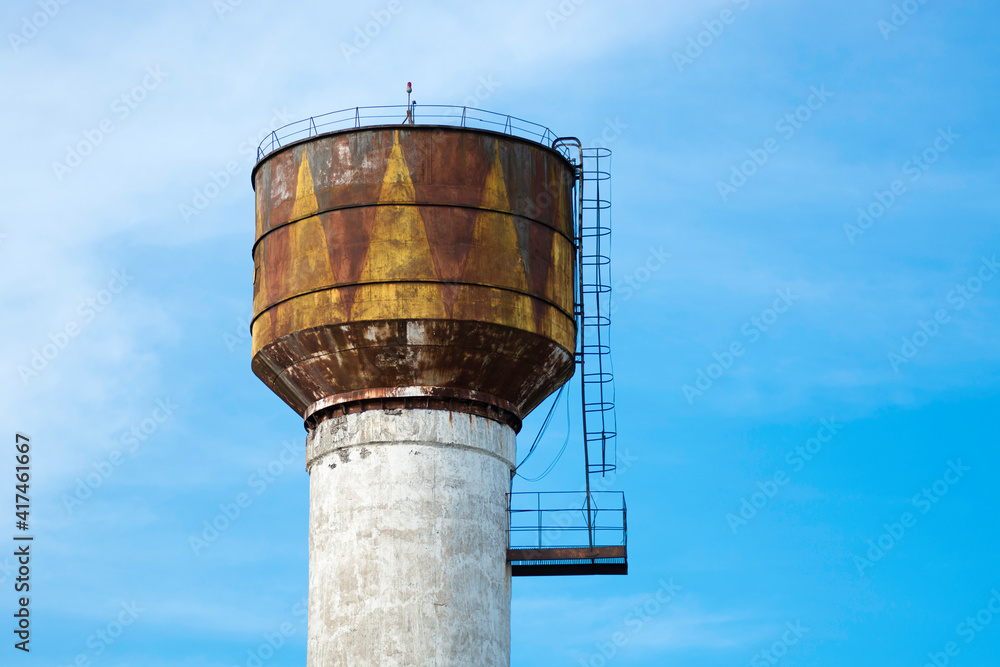 White with yellow brown color rusted old water tower on the background of a bright blue spring sky. With cornice and stairs.