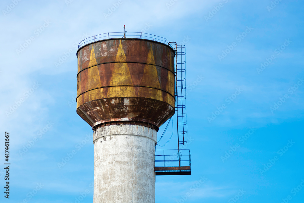 White with yellow brown color rusted old water tower on the background of a bright blue spring sky. With cornice and stairs. White with yellow brown color rusted old water tower on the background of a bright blue spring sky. With cornice and stairs.