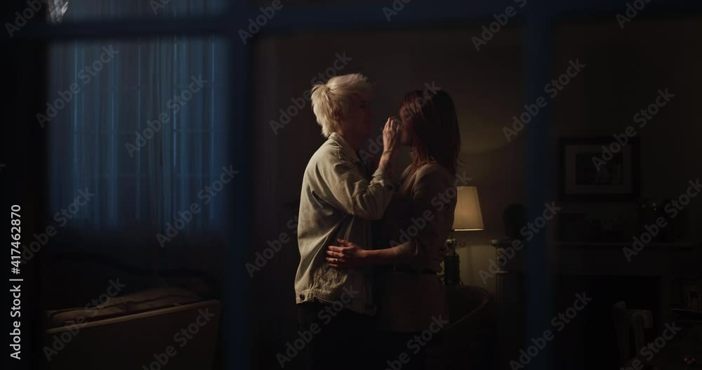 Cinematic shot from window of young happy homosexual female gay and transgender man couple in love enjoying to embrace and kiss while dancing together and listening to the music at home at evening. Cinematic shot from window of young happy homosexual female gay and transgender man couple in love enjoying to embrace and kiss while dancing together and listening to the music at home at evening.
