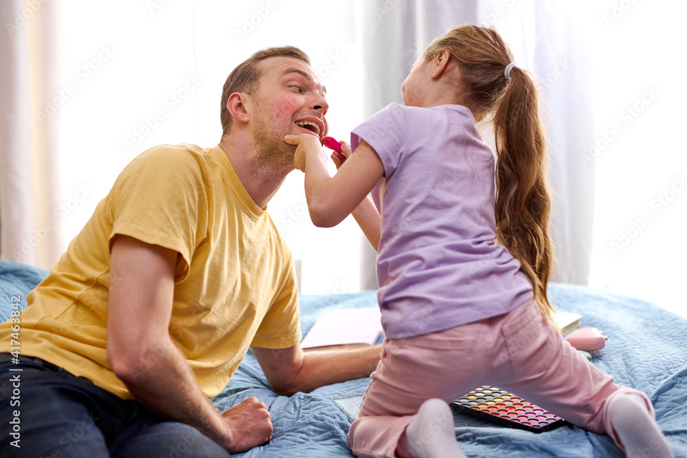 Dad and his child daughter are playing at home sitting on bed. Cute girl apply cosmetics on dad's face, do make-up. Family holiday and togetherness.