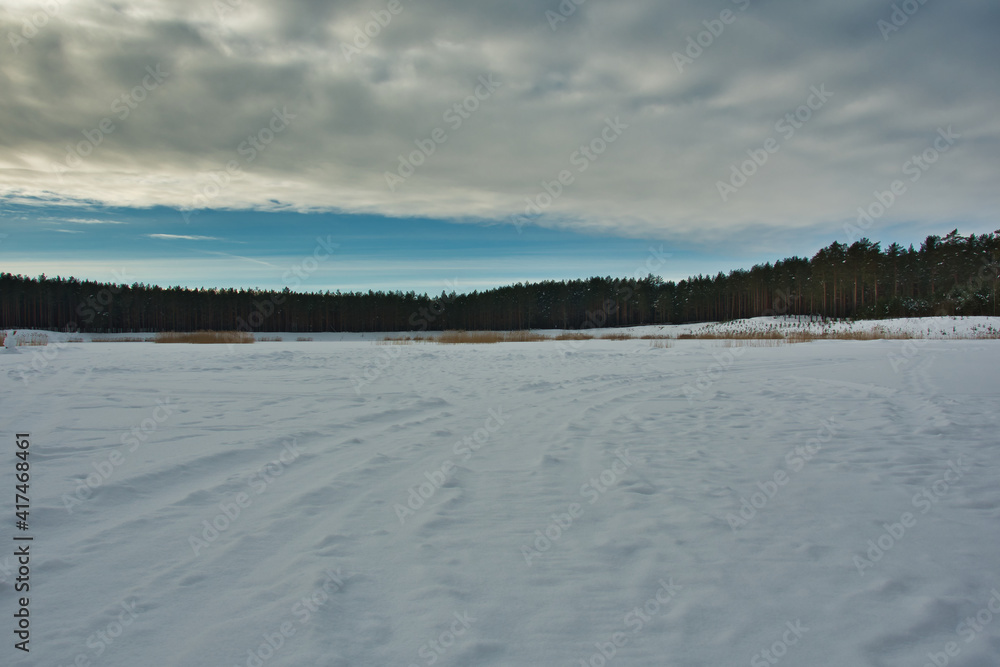 A landscape of a frozen snow-covered lake in a coniferous forest with yellow reeds on the edge, with the sky half-covered with clouds through which the winter sun breaks through.