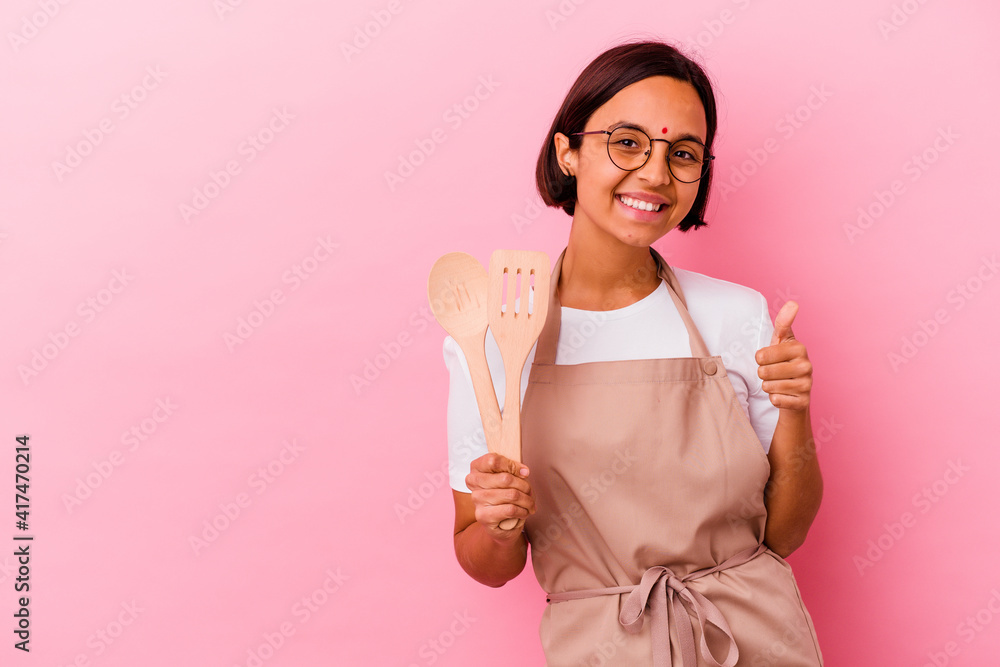 Young indian cook woman holding a wooden spoon and fork isolated smiling and raising thumb up Young indian cook woman holding a wooden spoon and fork isolated smiling and raising thumb up