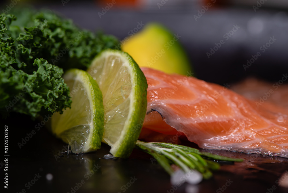 The Salmon Slices Served On A Black Ceramic Plate With Kale Leaf, Avocado, Lime Wedges, Rosemary Sprig And Coarse Tibetan Salt. Organic Food. Side View. Serving Example. Selective Focus. The Salmon Slices Served On A Black Ceramic Plate With Kale Leaf, Avocado, Lime Wedges, Rosemary Sprig And Coarse Tibetan Salt. Organic Food. Side View. Serving Example. Selective Focus.
