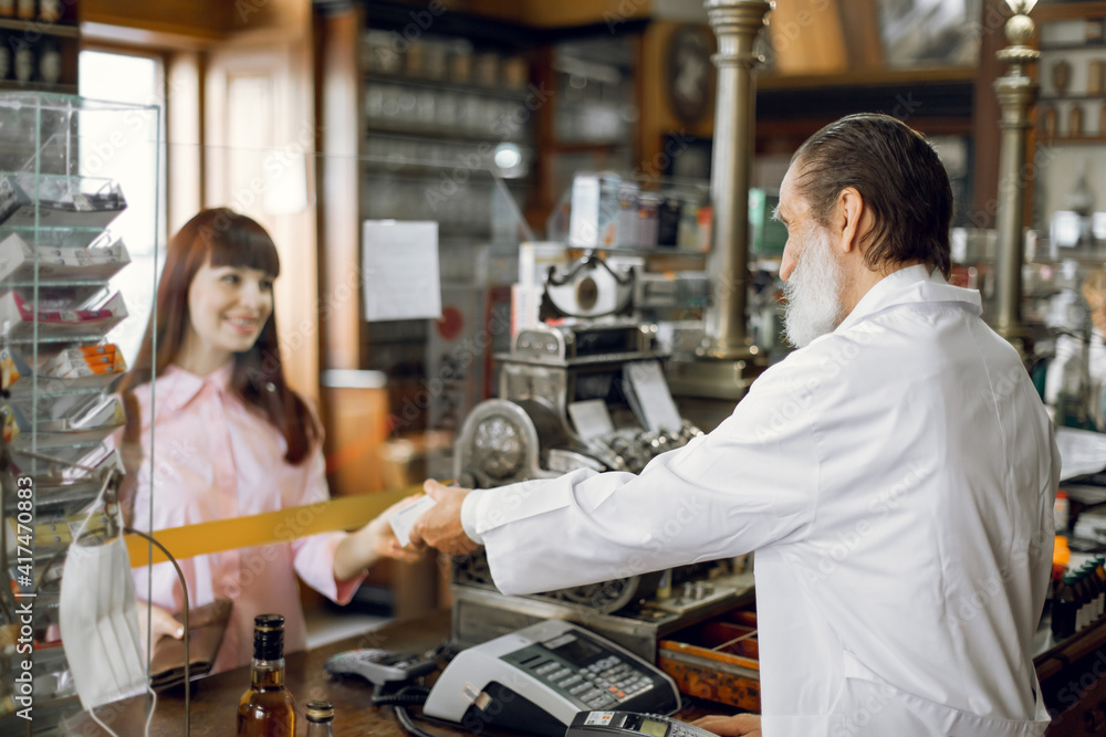 Back view of professional high-skilled mature man pharmacist selling medicines to young beautiful Caucasian woman patient. Drug dispensing at ancient old pharmacy.