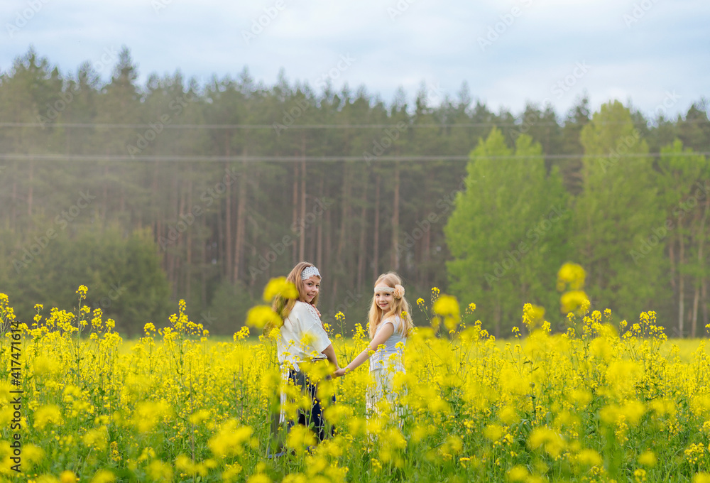 Two girls are walking along a blooming rapeseed field. Children play in a field with yellow flowers. Beautiful yellow field against the background of the forest. Hold hands