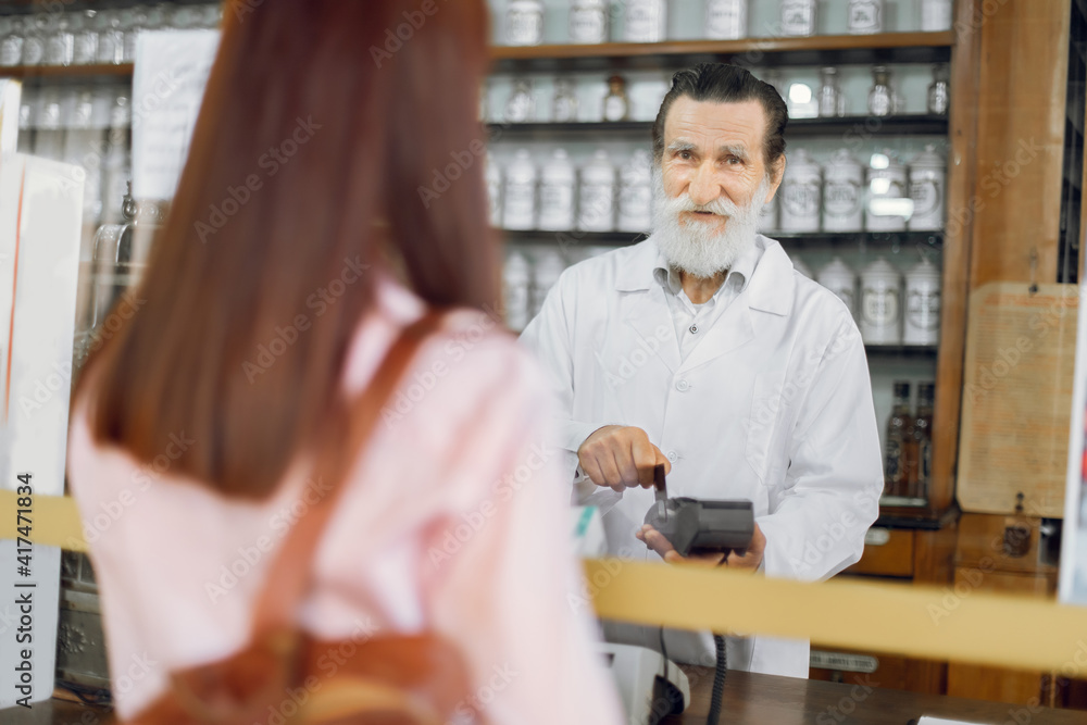 Back view of young Caucasian woman customer, paying for pills with credit card in old vintage pharmacy. Smiling senior man pharmacist swiping the credit card through payment terminal. Back view of young Caucasian woman customer, paying for pills with credit card in old vintage pharmacy. Smiling senior man pharmacist swiping the credit card through payment terminal.