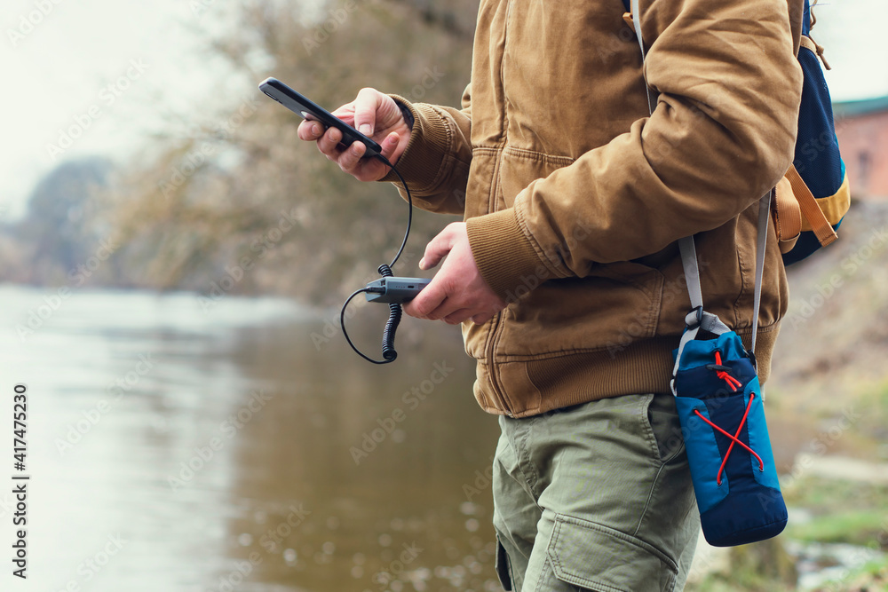 Fototapeta premium Tourist holds a portable charger with a smartphone in his hand. A man on the background of a lake with a backpack charges the telephone with Power Bank. Concept on the theme of tourism.