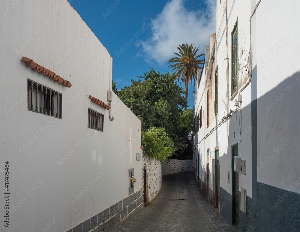 Narrow street in Agaete city center with traditional architecture, old white houses in colonial style, stone wall and tropical garden with palm trees. Gran Canaria, Canary Islands, Spain