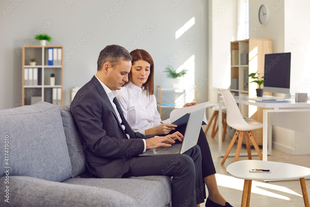 Two serious focused business people sitting on couch while meeting in the office. Man and woman working with data together, using laptop computer, doing paperwork