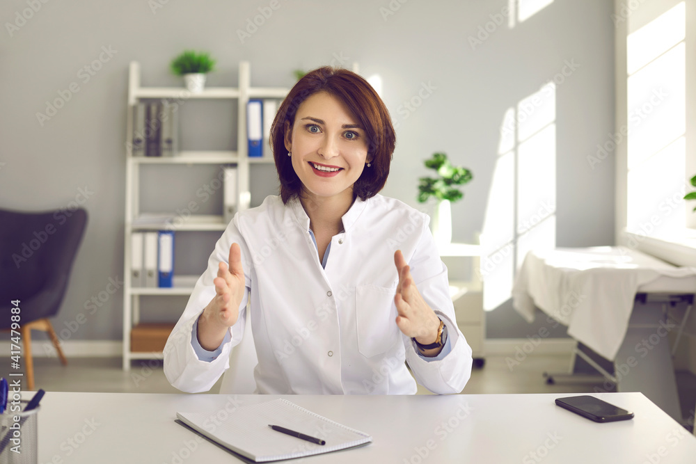Portrait of professional doctor talking to patient sitting at desk in medical office. Happy woman who works as general practitioner at hospital giving online consultation and smiling at camera Portrait of professional doctor talking to patient sitting at desk in medical office. Happy woman who works as general practitioner at hospital giving online consultation and smiling at camera