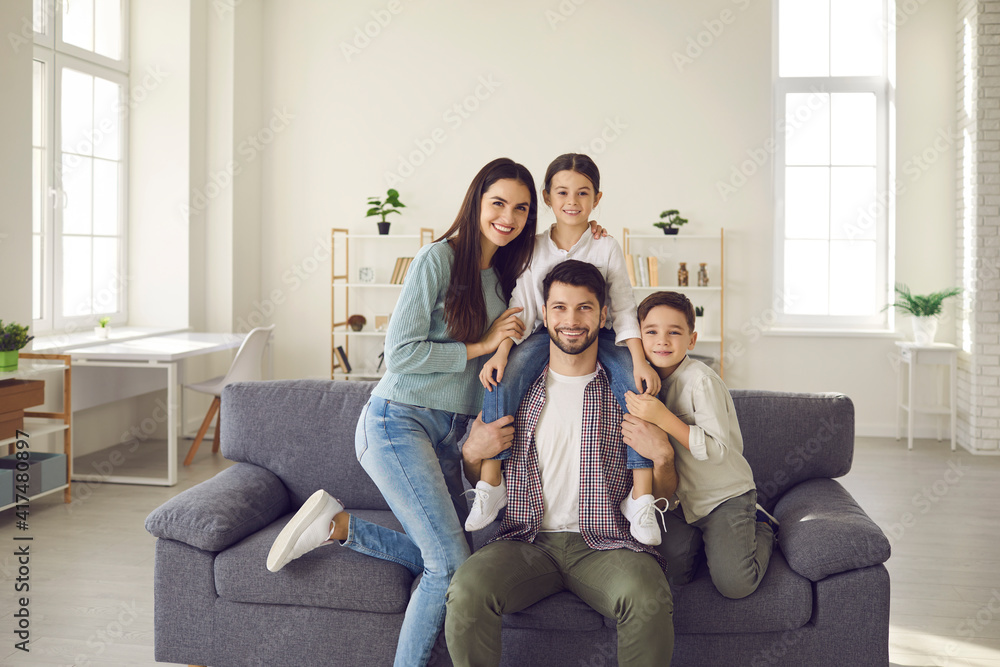 Portrait of happy young family with kids at home. Cheerful mother, father and little children smiling and looking at camera, sitting on sofa in the living-room of their new house or apartment