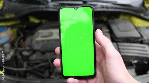 Young man holds a mobile smartphone in his hand and flips swipes right his finger on screen on background engine compartment of the car, car with an open hood