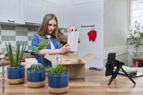 Woman recording video on smartphone unpacking cardboard box. Pet parrot helping to unpack