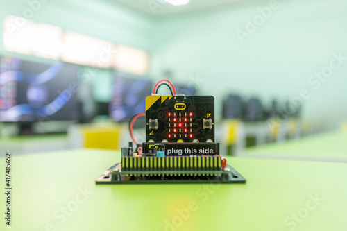 Microbit is a small circuit board, displaying a cross. It is set on a table in a computer science technology classroom.