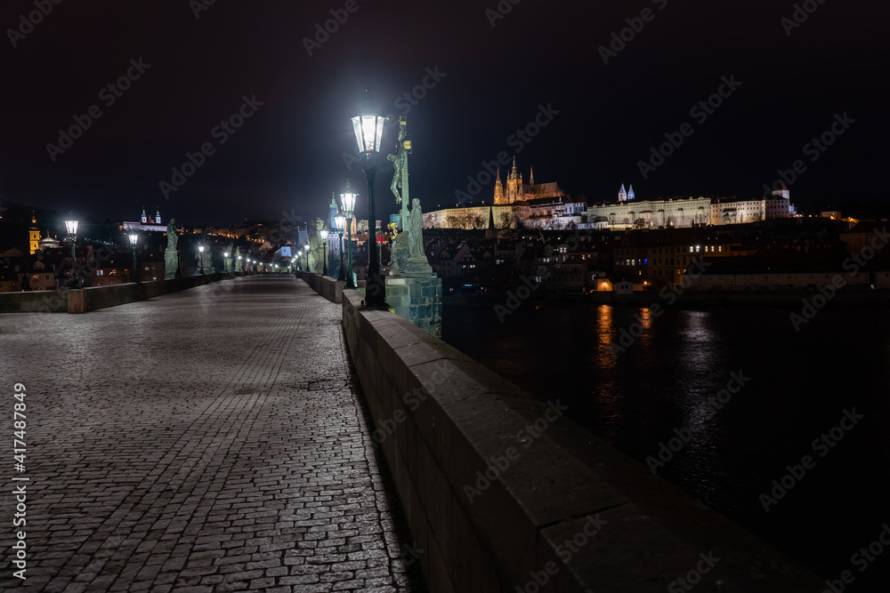 view of a stone pavement light from a lantern on Charles Bridge and illuminated statues and in the background the Church of St. Vitus and Prague Castle.
