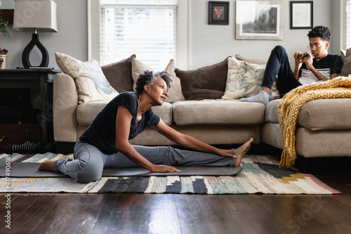 Black mom stretches in family room, teenage son on phone in background, fitness, health and wellness photo