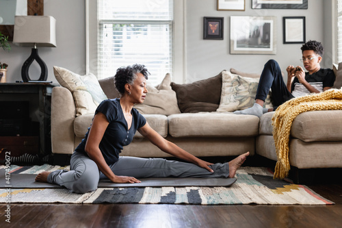 Black mom stretches in family room, teenage son on phone in background, fitness, health and wellness