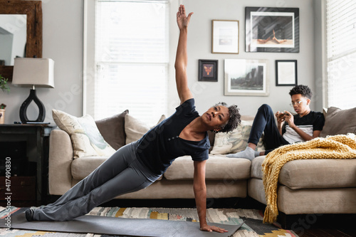 Black mom working out in family room, teenage son on phone in background, side plank exercise