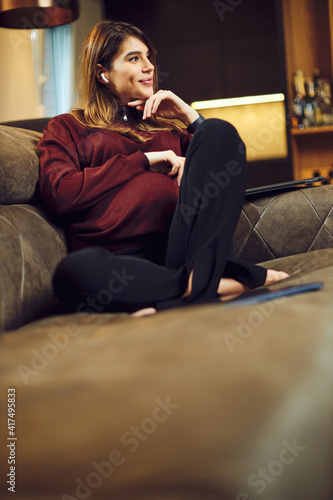 A pregnant woman sitting at home with laptop in lap, putting earphones and relaxing.