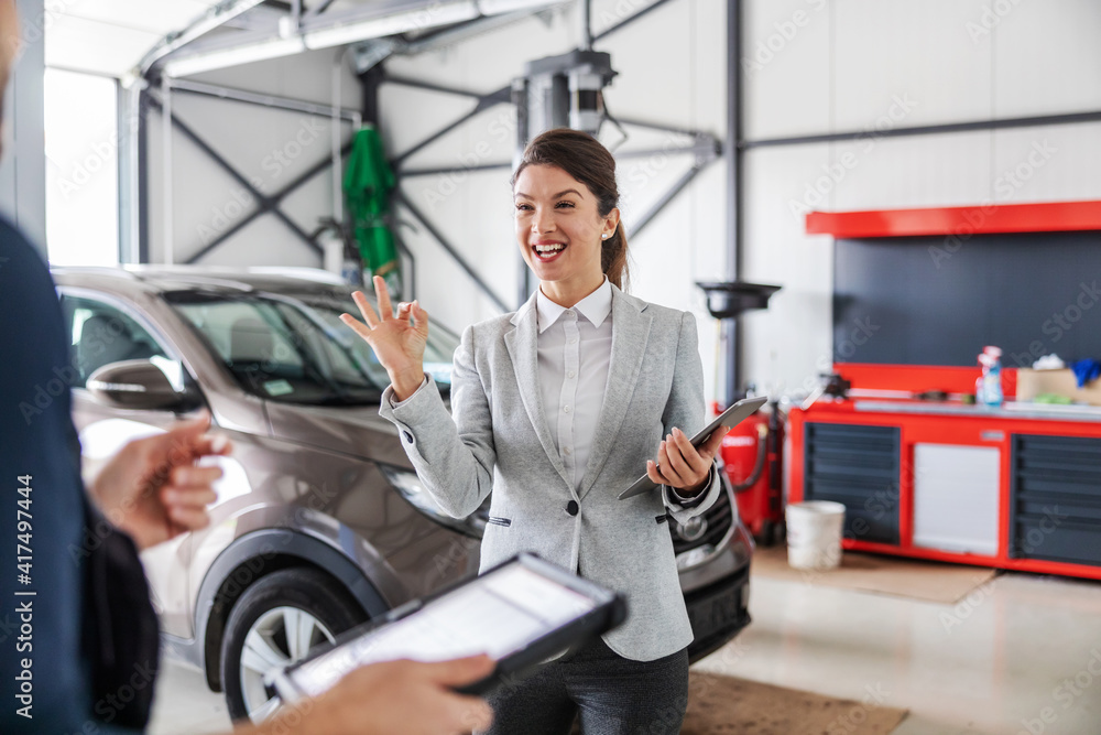 Smiling friendly car seller talking to a mechanic and showing okay hand gesture while standing in car salon. Smiling friendly car seller talking to a mechanic and showing okay hand gesture while standing in car salon.