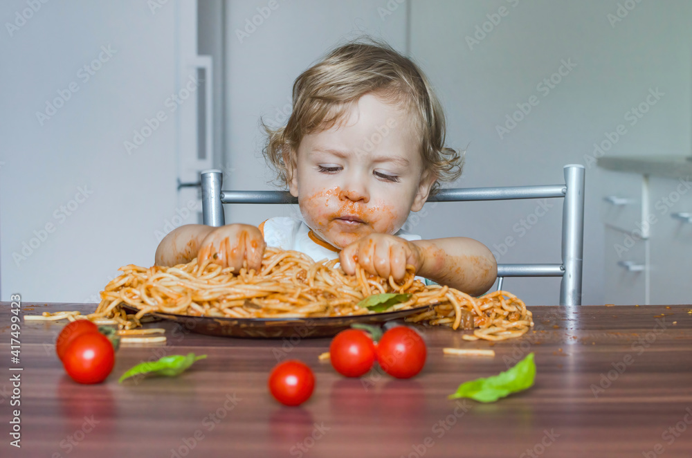 Funny baby child getting messy eating spaghetti with tomato sauce from a large plate, by itself with his hands, at home Funny baby child getting messy eating spaghetti with tomato sauce from a large plate, by itself with his hands, at home