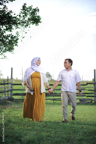 Affectionate young Asian Muslim couple in love wait for child, walk and hold together on green grass, have positive expression, want to become parents soon.