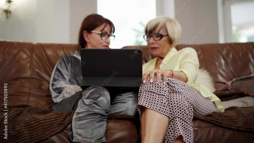Tablet on female knees with two blurred confident stylish senior women sitting on couch and talking at background. Fashionable Caucasian retirees in eyeglasses surfing Internet at home indoors. Tablet on female knees with two blurred confident stylish senior women sitting on couch and talking at background. Fashionable Caucasian retirees in eyeglasses surfing Internet at home indoors.