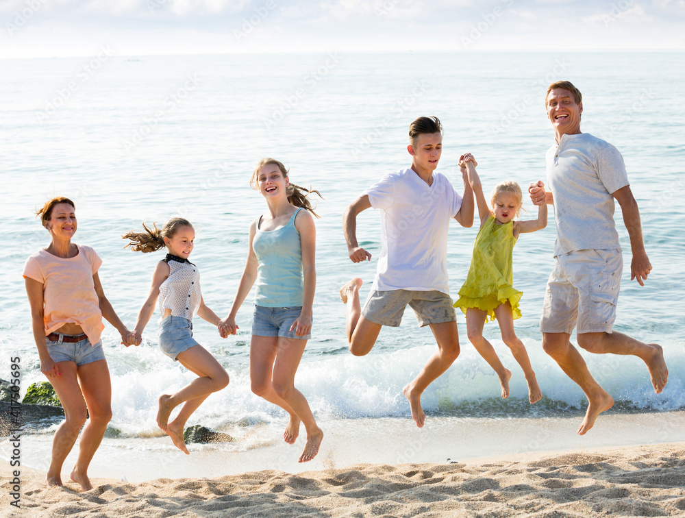 smiling man and woman with four children holding hands and jumping on sandy beach .