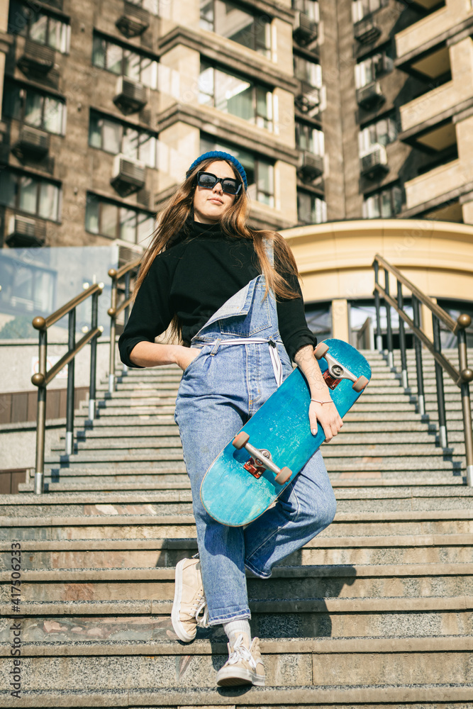 Stylishly dressed woman in blue denim jumpsuit posing with skateboard. Street photo. Portrait of girl holding skateboard. Lifestyle, youth concept. Leisure, hobby and skate in the city