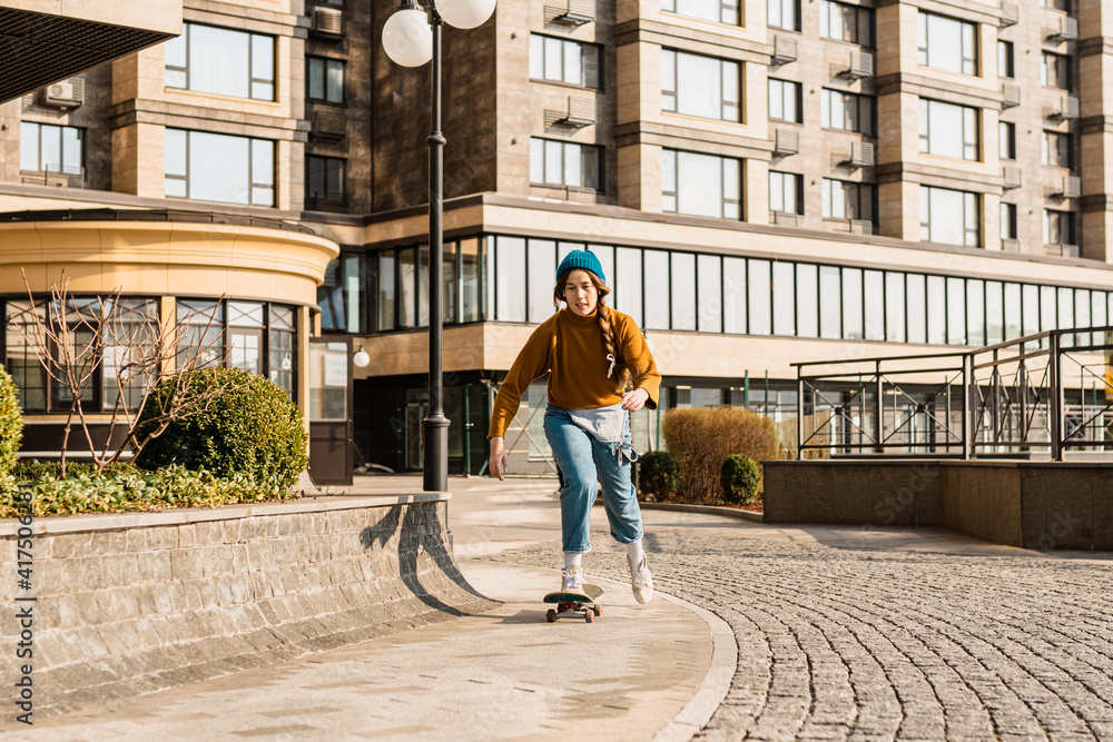 Fototapeta premium Young handsome woman riding on a skateboard. Portrait of sport female model in urban style. Skateboarding at city. Street style outfit. Urban woman ride on skate. Hipster girl on skateboard