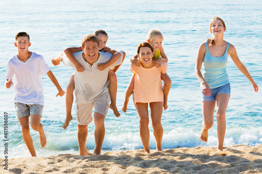 Large cheerful family of six people happily running and carrying kids on parents back on beach