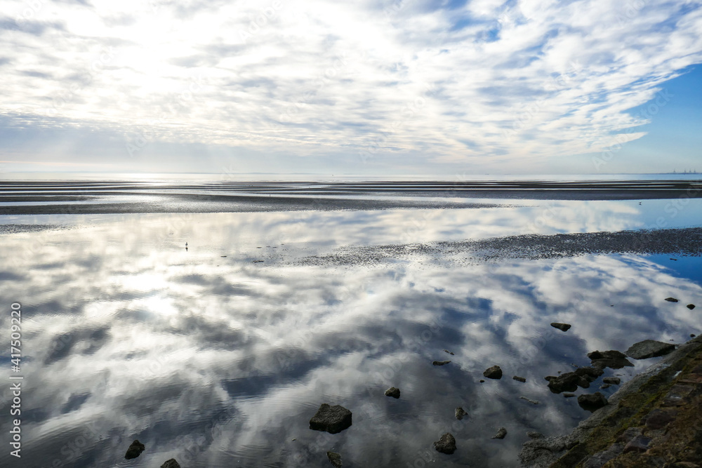 Sandgate at low tide. Sand with blue, cloudy sky with reflection of sky in water. Rocks at the bottom of the photo. 