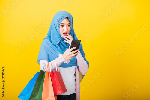 Asian Muslim Arab, Portrait of happy beautiful young woman Islam religious wear veil hijab funny she surprise on smartphone on hand and hold shopping bags, studio shot isolated on yellow background