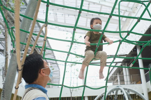 Cute little Asian boy child wearing washable face mask climb on jungle gym at pulbic indoor playground with his father during covid-19 outbreak, New normal lifestyle concept