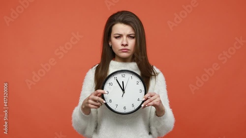 Displeased impatient woman in white sweater pointing at wall clock display, asking you to hurry up, warning about time out and deadline, punctuality. Indoor studio shot isolated on orange background