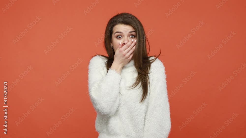 I will not say anything. Extremely shocked surprised brunette woman in white sweater covering her mouth with hand, looking at camera with big eyes. Indoor studio shot isolated on orange background I will not say anything. Extremely shocked surprised brunette woman in white sweater covering her mouth with hand, looking at camera with big eyes. Indoor studio shot isolated on orange background