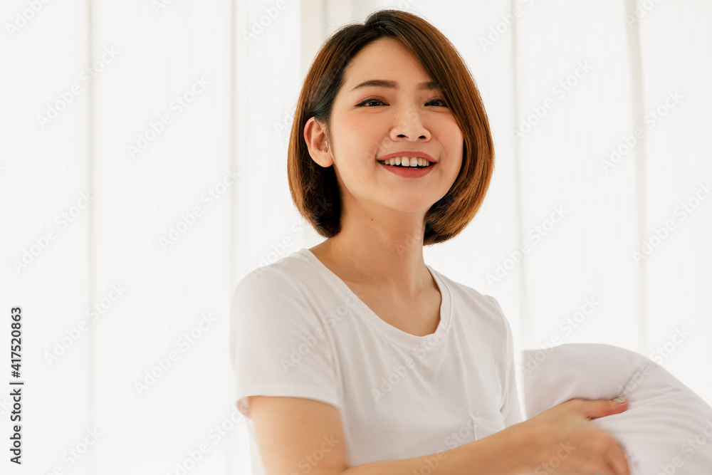 Adult beautiful asian woman pretty with short brown hair and wearing white t-shirt holding white pillow smiling and happy casual in morning on one day Adult beautiful asian woman pretty with short brown hair and wearing white t-shirt holding white pillow smiling and happy casual in morning on one day